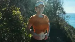 Frau in orangenem Shirt zieht Leggings runter und zeigt blanken Po am sonnigen Klippenrand über dem Meer