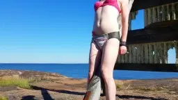 Frau in pinkem BH und nasser Windel steht gefesselt an Holzpfosten am sonnigen Meer